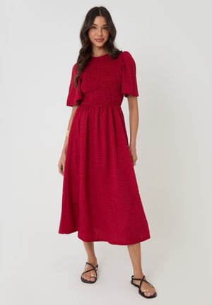 Femme portant une robe midi rouge à manches bouffantes et corsage froncé, debout dans une posture détendue avec des sandales plates noires sur un fond blanc.
