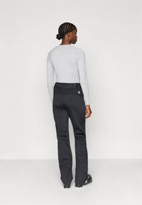 Γκρι μακρυμάνικη μπλούζα συνδυασμένη με μαύρο παντελόνι που διαθέτει υφασμένο ζωνάρι και φερμουάρ στους αστραγάλους. Το μοντέλο είναι γυρισμένο πλάτη στην κάμερα.