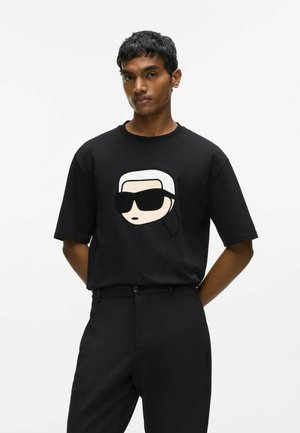 Mann trägt ein schwarzes T-Shirt mit großem Comic-Gesicht mit Sonnenbrille auf der Vorderseite, kombiniert mit schwarzen Hosen.