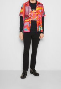 Chemise colorée à manches courtes avec des motifs abstraits en rose, bleu et jaune ; portée par-dessus un t-shirt noir à manches longues et un pantalon noir, avec des chaussures noires.