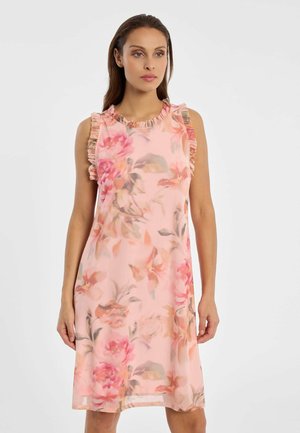 Frau in ärmellosem, rosa Blumenkleid mit Rüschenbesatz an Schultern und Ausschnitt, stehend vor schlichtem weißem Hintergrund.