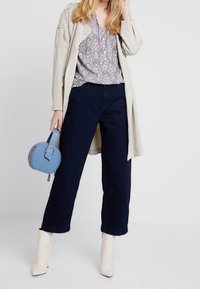 Jasnobeżowy płaszcz typu trencz na kwiecistej bluzce, w połączeniu z ciemnoniebieskimi, szerokimi spodniami dżinsowymi i kremowymi botkami za kostkę. Trzyma małą, niebieską okrągłą torebkę.