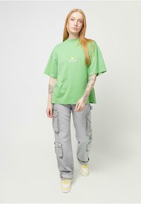Zielona, oversize'owa koszulka z graficznym logo i krótkimi rękawami, zestawiona z jasnoszarymi spodniami cargo z wieloma kieszeniami i luźnym krojem.