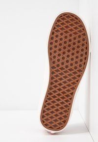 Baskets de couleur claire avec une semelle en caoutchouc marron texturée à motif géométrique. Vue latérale montrant une tige lisse et un style minimaliste.