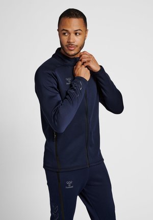 Mand iført marineblå sportjakke og bukser, der lynlåser jakken, mens han kigger til siden mod en ensfarvet baggrund.