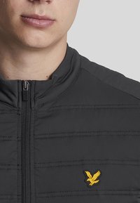 Giacca nera con zip in tessuto texture, caratterizzata da cuciture orizzontali e un piccolo logo a forma di uccello giallo sul lato sinistro del petto.