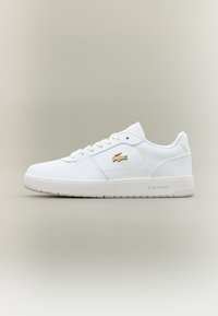 Bílé nízké tenisky s zlatým krokodýlem Lacoste, bílé tkaničky a mírně texturovaná podešev na neutrálním pozadí.