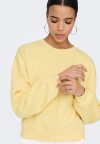 Vrouw met donker haar in een geel sweatshirt, met grote oorbellen en ringen, die omlaag kijkt terwijl ze haar mouw aanpast tegen een effen achtergrond.