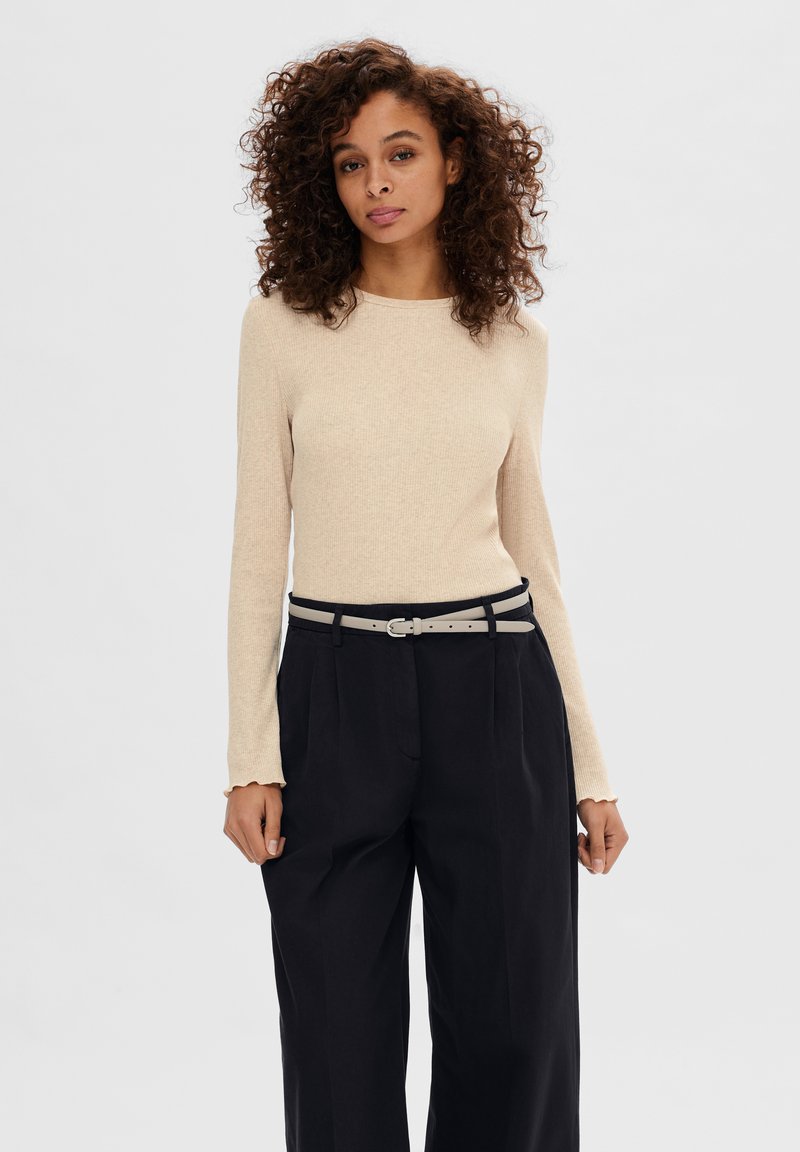 Top lungo beige a coste con polsini a scalloped, abbinato a pantaloni neri a vita alta e una cintura grigia chiara.