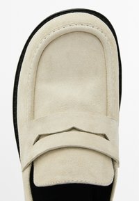Beige suede slip-on shoe with a rounded toe and outlined saddle stitch detailing. Black rubber sole, smooth texture, and minimal design.