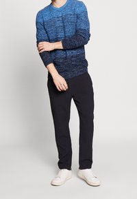 Pull en tricot dégradé bleu avec manches longues, col rond, tissu texturé, associé à un pantalon noir et des baskets blanches.