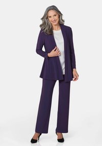 Lila langärmlige Jacke über einem weißen Oberteil, kombiniert mit passenden lila Hosen. Glattes Material, offenes Frontdesign und schlichtes Schuhwerk.