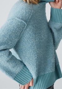 Personne portant un pull en laine bleu clair texturé avec des poignets et un ourlet côtelés épais, ajustant le col d'une main.