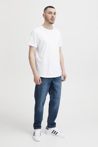 Fehér háttér előtt álló férfi, sima fehér pólót, kék farmert és fekete csíkos fehér sportcipőt visel.