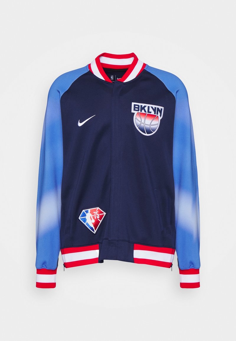 Veste bleu marine avec des manches en dégradé de bleu et rouge. Comprend le logo "BKLYN", le swoosh Nike et un écusson en forme de diamant. Col et poignets côtelés.