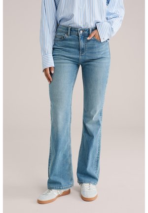 Személy világoskék csíkos ingben, magasderekú világoskék trapéznadrágban, fehér tornacipőben, egyik kezét a zsebében tartva.