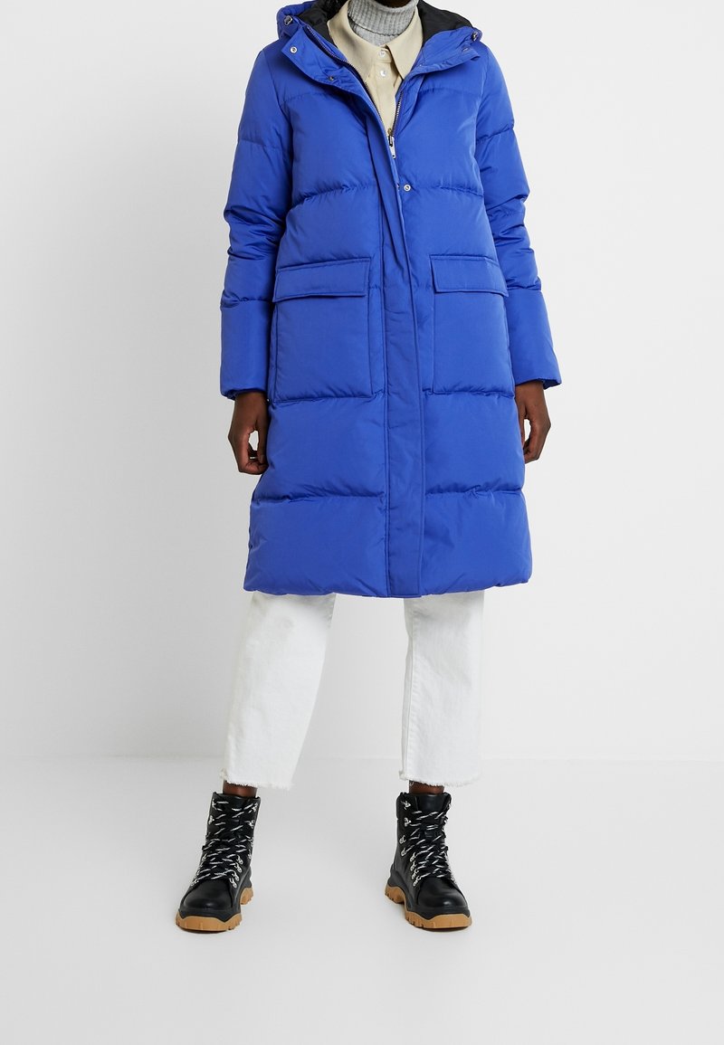 Blue puffer coat with a hood, front zip, and two large pockets; features a quilted design and is paired with black lace-up boots and white pants.