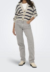 Femme portant un gilet beige à rayures, un haut blanc, un jean gris clair et des bottines crème, debout avec une main dans la poche.