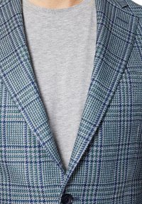 Płaszcz w kratkę w odcieniach niebieskiego i zieleni, wykonany z teksturowanej tkaniny. Posiada klapę oraz zapięcie na jeden guzik, noszony na szarym koszuli.