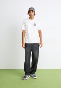 Hvid kortærmet t-shirt med et cirkulært logo på brystet, parret med mørkegrå jeans og grå sneakers. Neutral baggrund med grøn gulv.