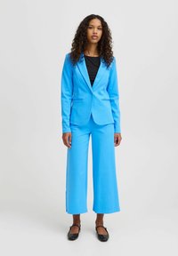 Felblauw pak met een nauwsluitende blazer en wijde broekspijpen, gemaakt van een gladde stof. De blazer heeft subtiele voorzakken en een knoop.