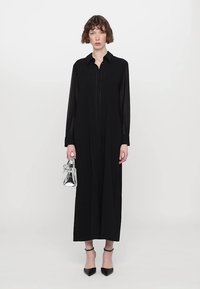 Vestido negro de manga larga con cuello de camisa y detalles de costura vertical, acompañado de un bolso transparente y unos tacones negros de punta.
