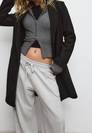 Une femme porte un pull gris côtelé court à fermeture éclair sur un haut noir, un pantalon de survêtement gris clair à cordon et un manteau long noir, posant avec une main dans la poche.