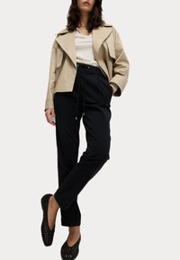 Beige ytterjacka med breda slag, i kombination med svarta justerbara dragsnoddbyxor och svarta vävda slip-on skor.