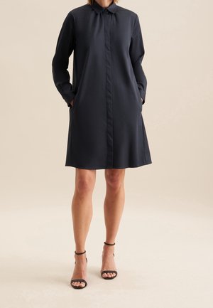 Frau trägt ein knielanges, langärmeliges schwarzes Hemdblusenkleid mit Kragen und verdeckten vorderen Knöpfen, kombiniert mit schwarzen Schuhen mit Knöchelriemen.