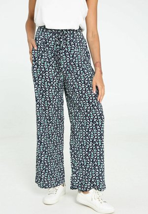 Kvinde iført løstsiddende sorte bukser med blå og hvidt blomsterprint og hvide sneakers, stående mod en ensfarvet baggrund.