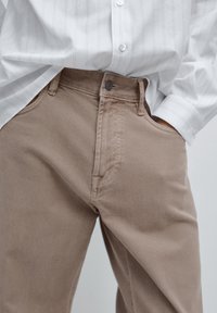 Beżowe dżinsy o prostym kroju, wyposażone w zapięcie na guzik i boczne kieszenie. Noszone pod białą koszulą w paski z długim rękawem.