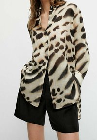 Chemise léopard à boutons, portée avec un short noir à mi-cuisses, mains dans les poches, présentée sur une personne devant un fond uni.