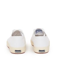 Scarpe slip-on bianche in tela con una suola in gomma beige testurizzata e un piccolo etichetta nera con logo sul tallone. Design semplice e minimale.