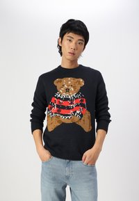 Zwarte trui met een gebreid teddybeerontwerp in het bruin, met een rood en zwart gestreepte sjaal. Lichtblauwe spijkerbroek eronder gedragen.