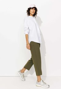 Une chemise blanche à boutons, un pantalon court olive et des baskets argentées. La tenue inclut une casquette rose pâle. Design minimaliste et moderne.