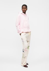 Žena nosící světle růžovou mikinu s kapucí a krémové široké kalhoty s vícebarevnými potisky koní, stojící s rukama v kapsách.