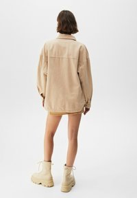 Veste en jean beige surdimensionnée avec des manches longues, des poignets à boutons et un ourlet en courbe ; associée à une mini-jupe beige et des bottes imposantes beiges.