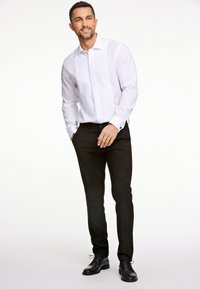 Camicia bianca a maniche lunghe con colletto rigido, abbinata a pantaloni neri e scarpe eleganti nere, in piedi su uno sfondo neutro.