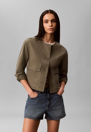 Frau trägt eine olivgrüne Jacke mit Knöpfen, großen Taschen und hochgekrempelten Ärmeln, kombiniert mit dunklen Jeansshorts, steht vor einem grauen Hintergrund.