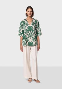 Blusa verde con motivo floreale, scollatura a V e maniche ampie, abbinata a pantaloni a gamba larga color crema e sandali bianchi con cinturini.