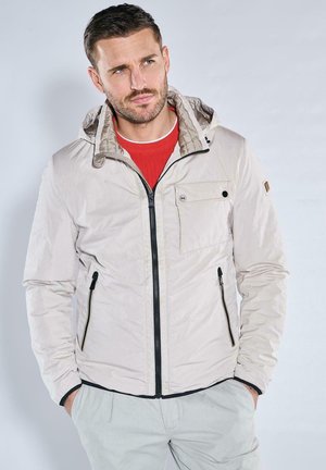 Light beige zip-up jacket with a hood, featuring a black zipper, front pocket, and textured interior. Worn over a red shirt.