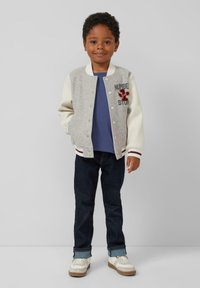 Grijs en crème kleurblok varsityjack met knopen, rode borduursels, over een blauwe shirt, gecombineerd met donkere jeans en witte sneakers.