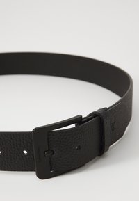 Cinturón de cuero negro con un acabado texturizado, que cuenta con una hebilla de metal negro y agujeros ajustables para el tamaño. Detalle sutil del logo en la correa.