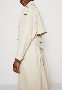 Mujer vestida con un abrigo trench beige con botones grandes y un cinturón atado en la parte de atrás, posando frente a un fondo claro y liso.