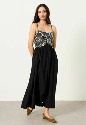 Frau in einem schwarzen ärmellosen Kleid mit großer weißer Blumenstickerei auf dem Oberteil, trägt olivgrüne spitze flache Schuhe, steht auf einem grauen Boden.