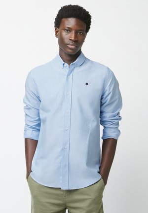 Jonge man met een lichtblauw overhemd met knopen en een klein boomlogo, handen in de zakken, tegen een effen witte achtergrond.