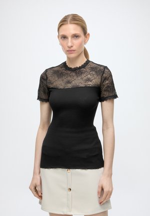 Femme portant un top noir moulant avec une partie supérieure et des manches en dentelle florale transparente, associée à une jupe beige boutonnée.