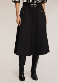 Falda midi negra con una textura suave, que presenta una cintura ceñida, bolsillos laterales y un detalle dorado brillante en el cinturón. Combinada con altas botas negras.