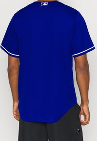 Královsky modrý baseballový dres s krátkým rukávem, bílými akcentními pruhy na rukávech a malým logem MLB na zadním výstřihu. Lehký materiál.