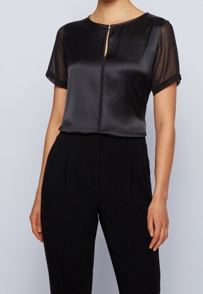 Svart satinblus med korta genomskinliga ärmar och detalj med nyckelhål i halsringningen, kombinerad med skräddarsydda svarta byxor. Slät textur och klassisk design.